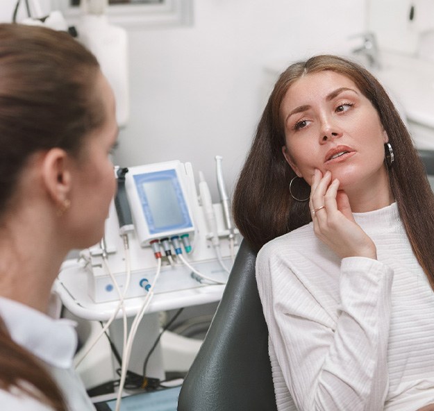 Woman in dental chair looking at dentist touching hand to jaw in pain