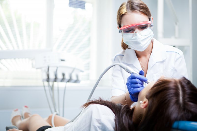 Woman wearing safety glasses and face mask using dental equipment to clean woman patient's teeth